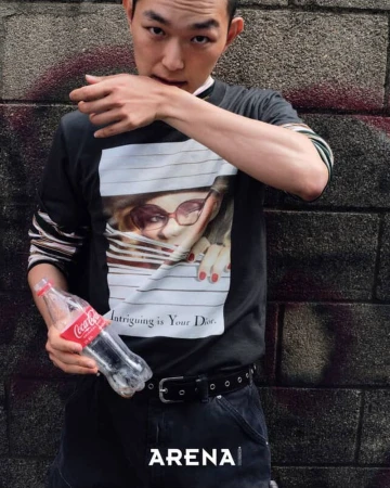 Asian male in black graphic tee, holding Coca-Cola bottle against graffiti wall. Urban fashion shot for magazine, casual style.