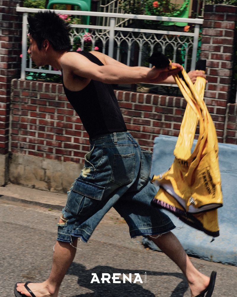 Athletic man runs outdoors wearing tank top and denim shorts, carrying yellow garment, showcasing active lifestyle and urban fashion.