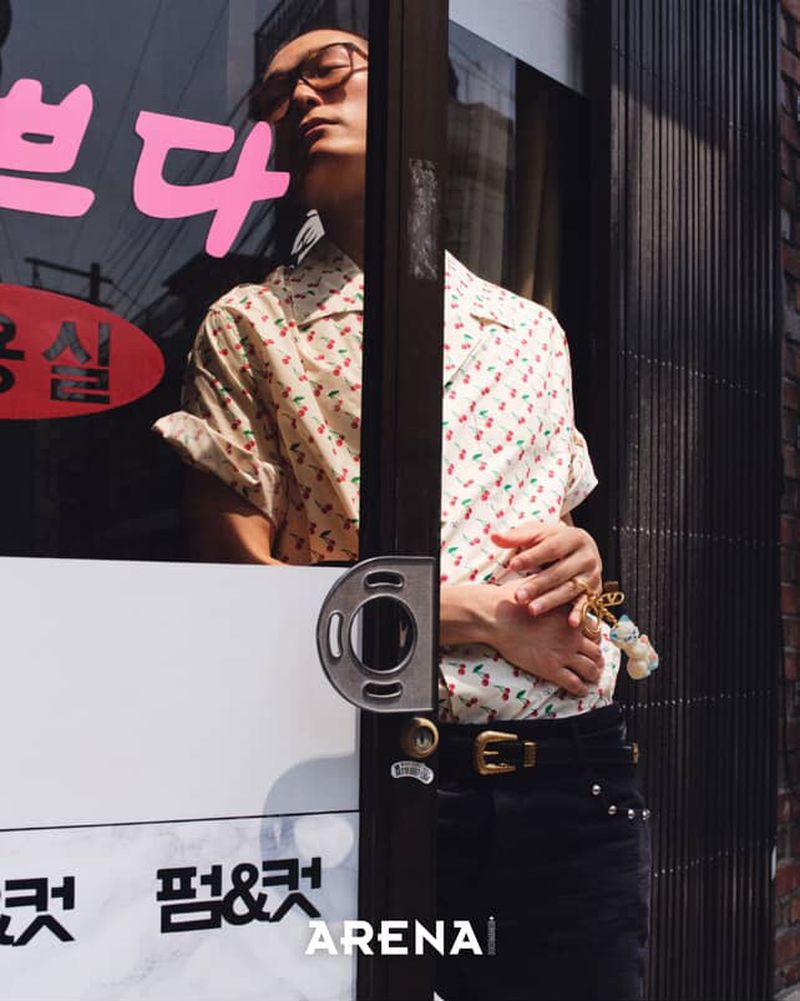 Stylish adult male poses in front of a storefront with patterned shirt and jeans. Eye glasses and jewelry are visible.