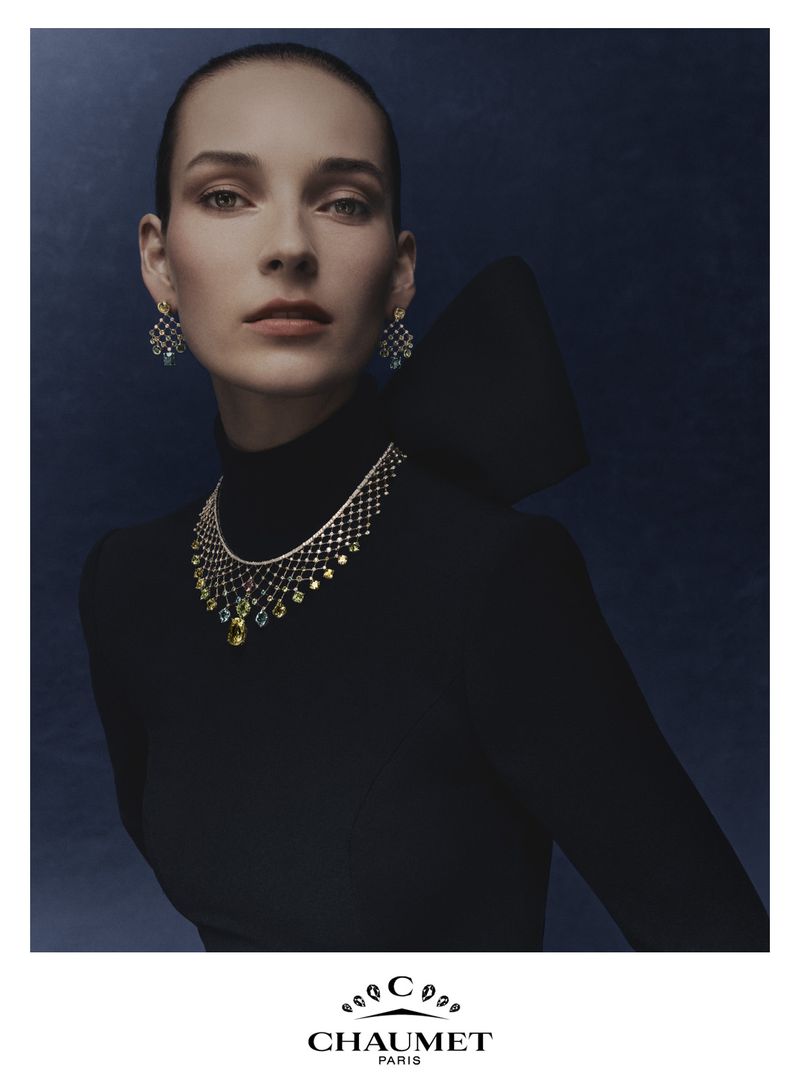 Model Julia Bergshoeff wearing a Chaumet gemstone statement necklace and drop earrings in a black high-neck gown against a blue background.