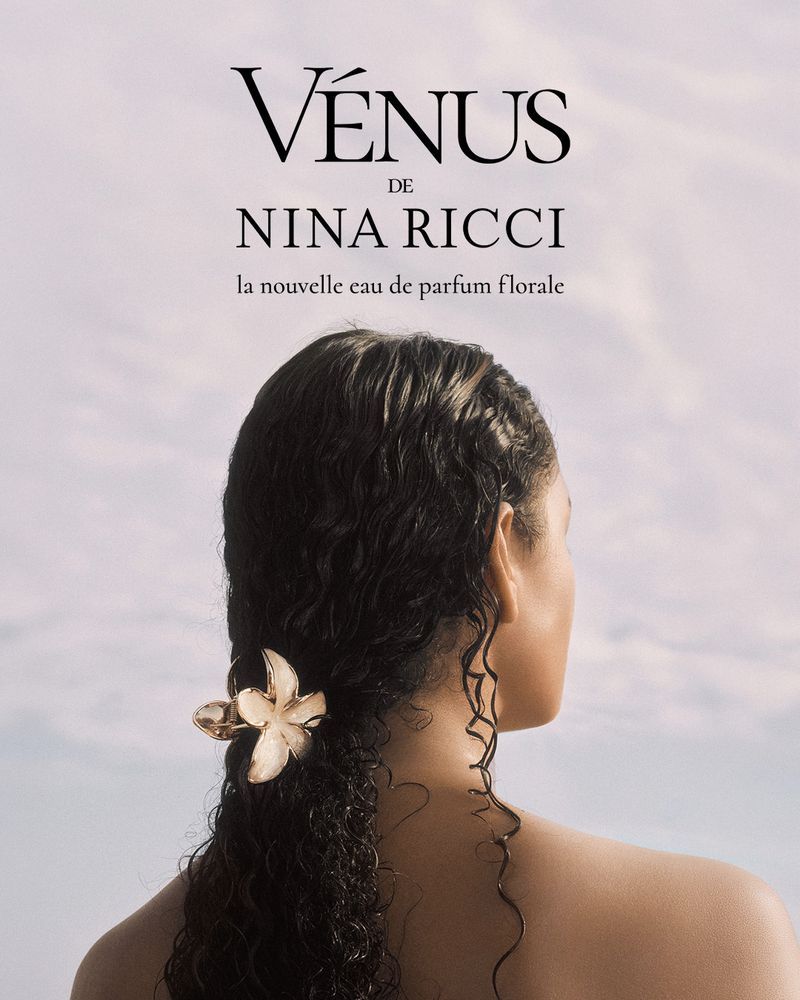 Model Tina Kunakey seen from behind with wet dark curly hair featuring a sculptural flower accessory against a pale sky.