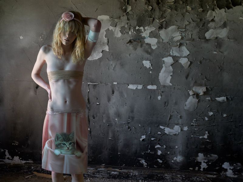 Artistic nude portrait of a young woman with blonde hair in a ruined interior. A kitten graphic is visible on her pink skirt.