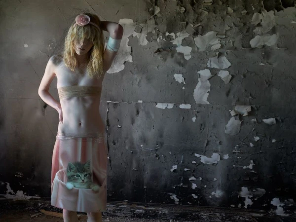 Artistic nude portrait of a young woman with blonde hair in a ruined interior. A kitten graphic is visible on her pink skirt.