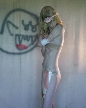 Artistic studio shot of a slender young adult female in a transparent garment and eye covering posing in front of graffiti art.