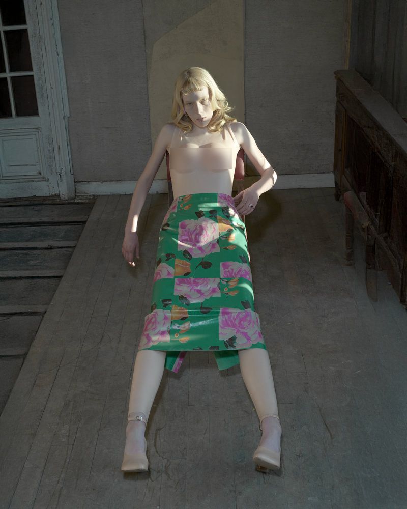 Woman poses wearing a floral skirt and nude top in a vintage setting with wood floors, walls, and a door, with neutral, soft lighting.