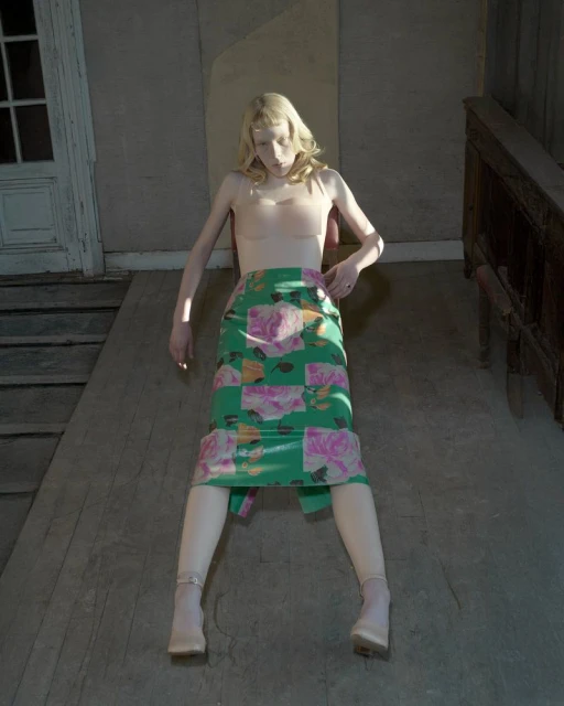Woman poses wearing a floral skirt and nude top in a vintage setting with wood floors, walls, and a door, with neutral, soft lighting.