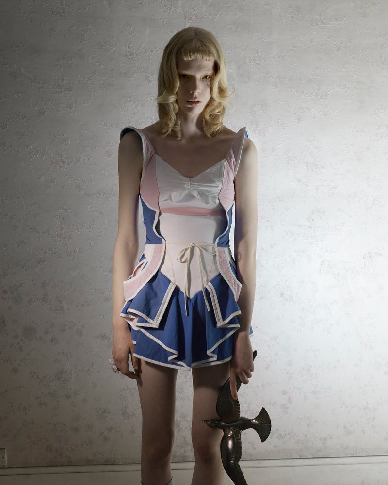A single fashion model poses indoors wearing a pink and blue garment and holding a bronze bird statue in a studio setting.