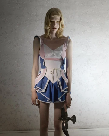 A single fashion model poses indoors wearing a pink and blue garment and holding a bronze bird statue in a studio setting.