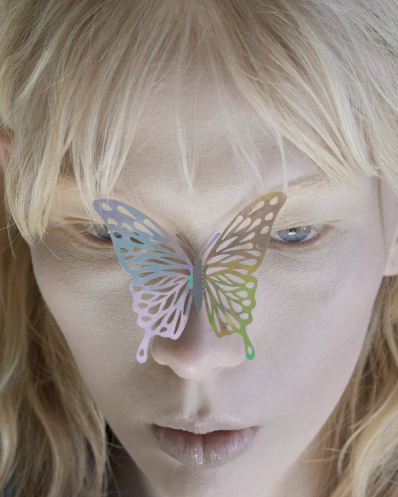 Close-up portrait of a fair-skinned woman with blonde hair, pale blue eyes, and a decorative butterfly on her face in an indoor setting.