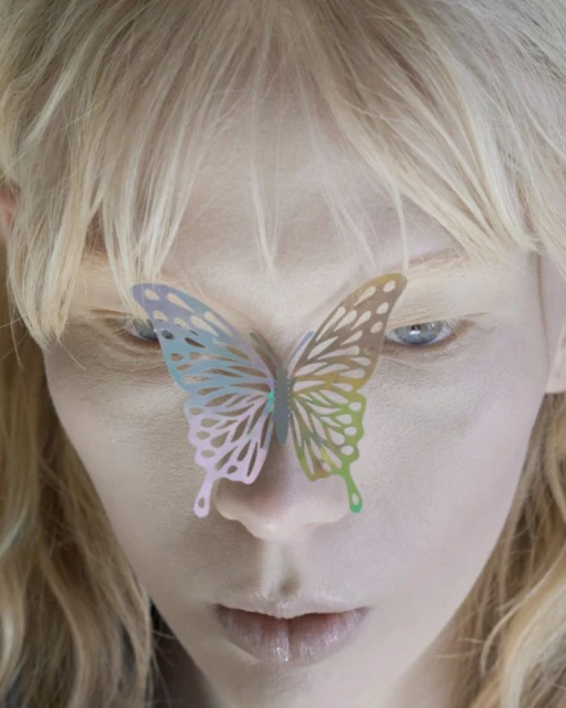 Close-up portrait of a fair-skinned woman with blonde hair, pale blue eyes, and a decorative butterfly on her face in an indoor setting.
