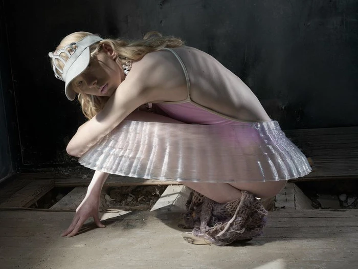 Model in a white visor and translucent skirt crouching against a dark textured interior background