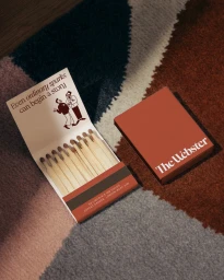 Branded terracotta matchbook open to reveal wooden matches alongside a closed matchbox for The Webster on a textured wool rug.