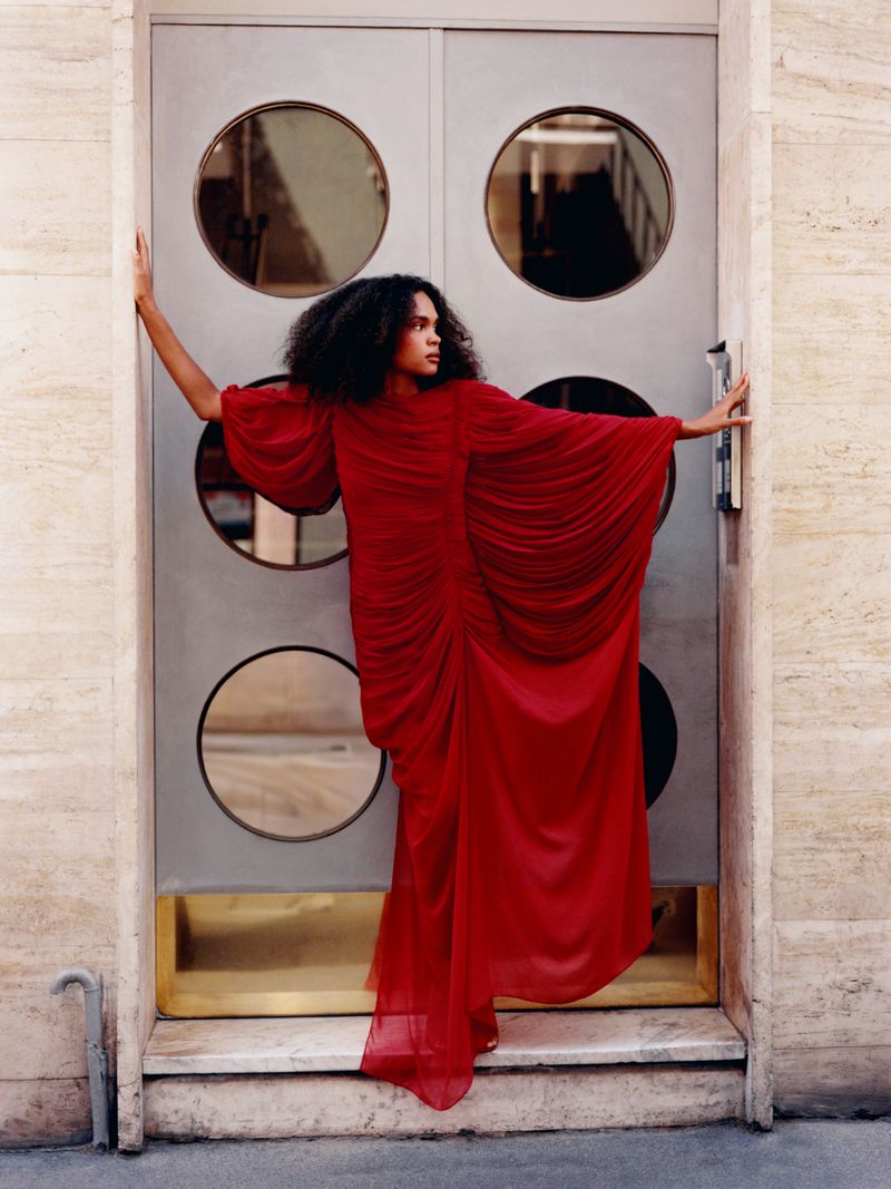 Fashionable adult in a long, draped red dress poses dramatically against a textured doorway with circular window details.