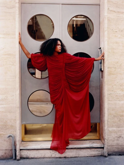 Fashionable adult in a long, draped red dress poses dramatically against a textured doorway with circular window details.