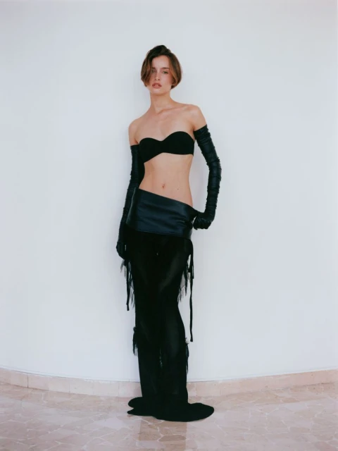 A slender woman poses against a white wall wearing a black strapless top, long gloves, and a black skirt with fringe details.
