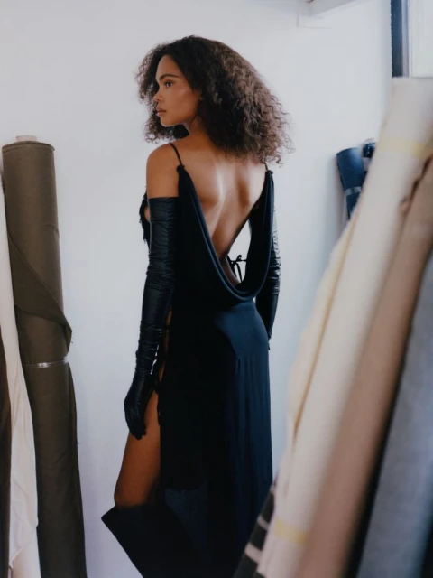 Back view of a model in a dark, low-back gown and long gloves, standing in a studio with rolls of canvas and white walls.