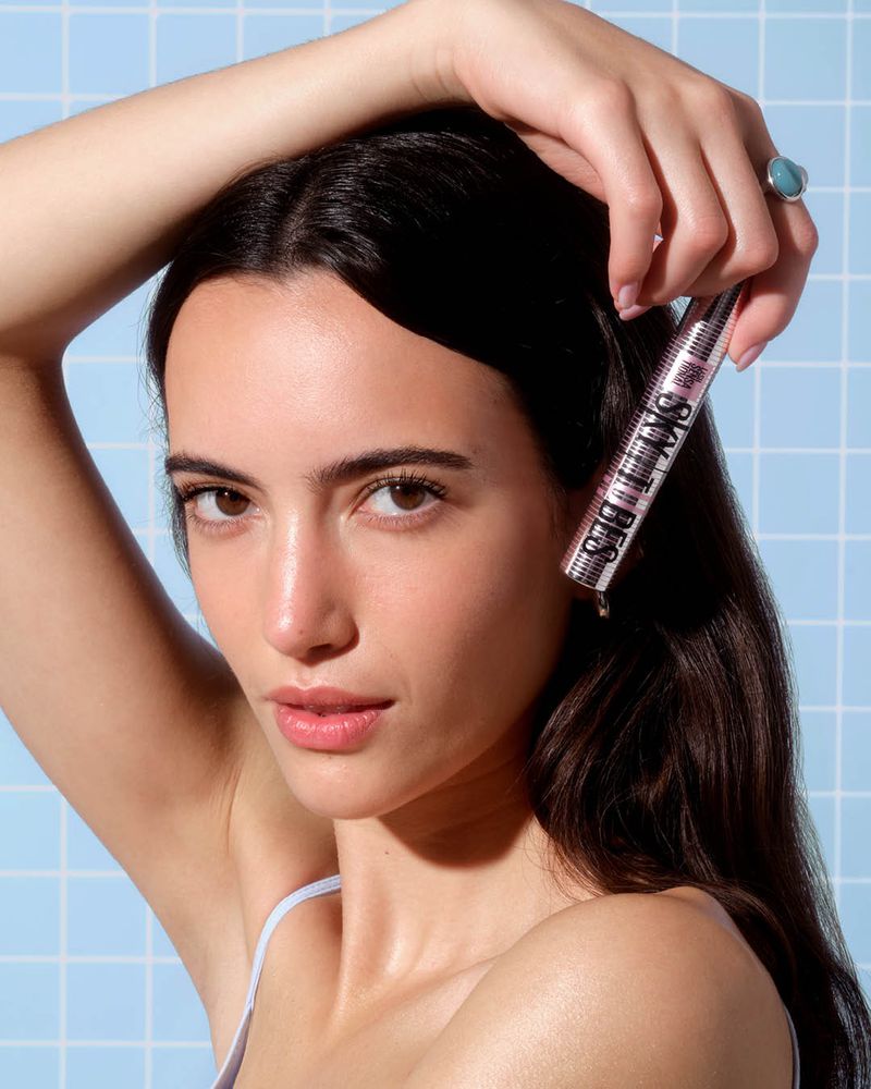 Close-up portrait of a woman holding a mascara tube against a light blue tiled background for ASOS.