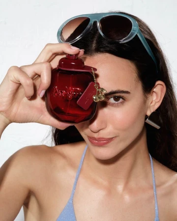 Woman in light blue sunglasses holding a deep red fragrance bottle over her eye against a plain white background.