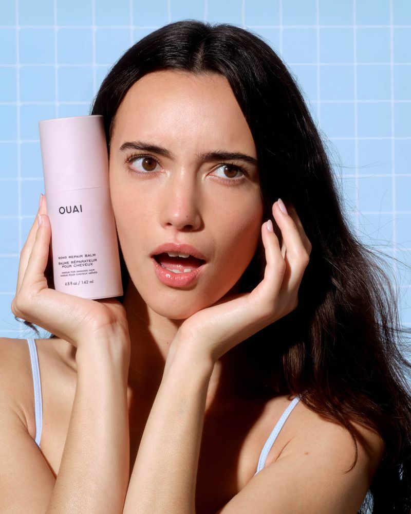 Model holding a pink OUAI hair repair balm bottle next to her face against a light blue tiled grid background.