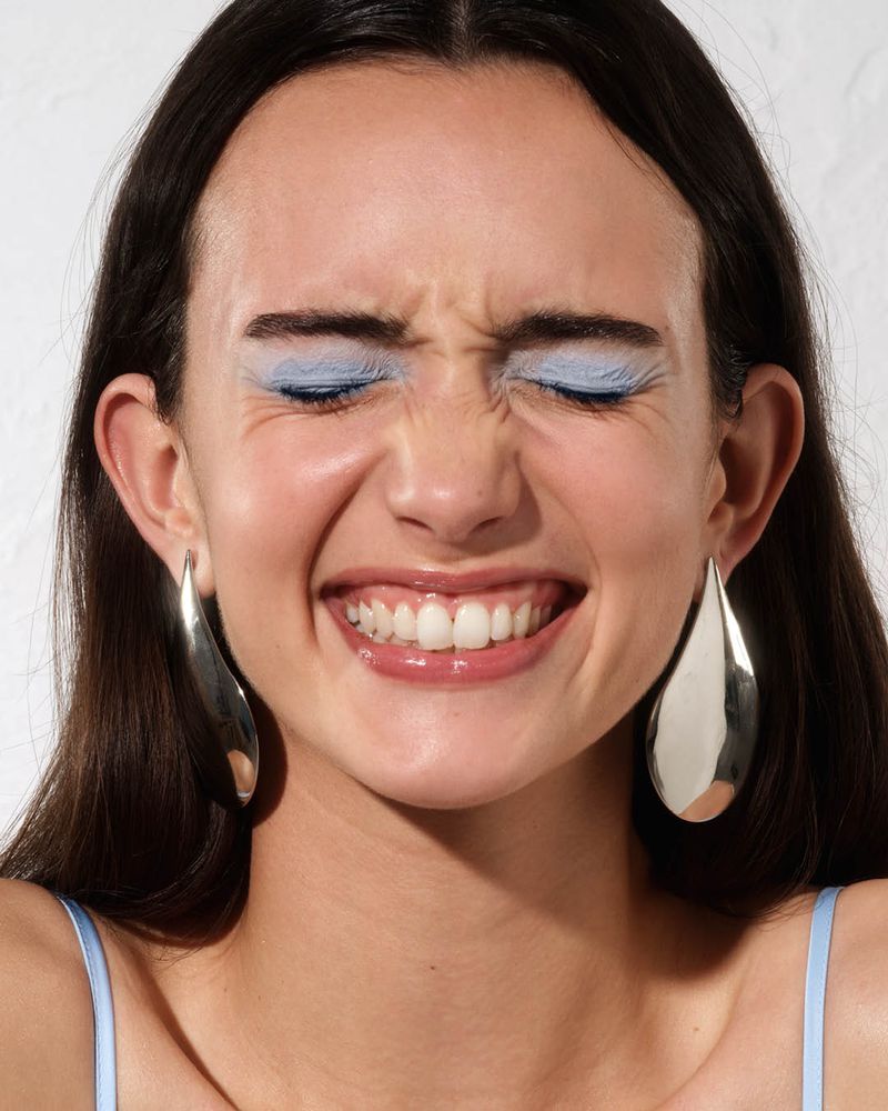 Close-up of a smiling woman with pastel blue eyeshadow and large polished silver teardrop earrings against a white backdrop.