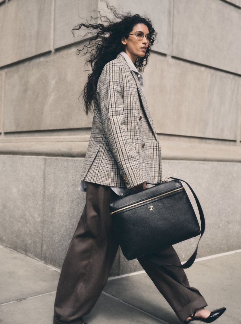 Model Jennifer Matias walking in a plaid blazer, wide brown trousers, and carrying a large black Chanel leather shoulder bag.