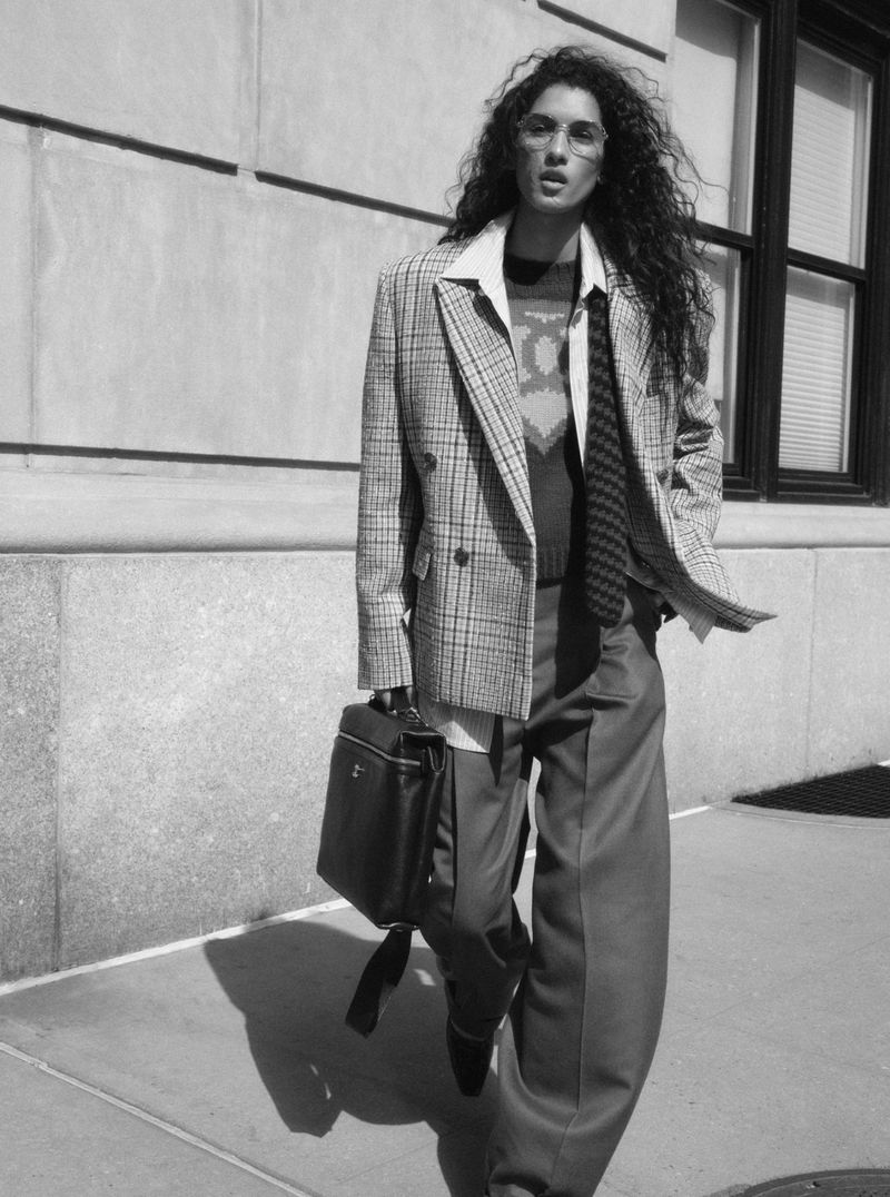 Model with long dark curly hair walking outdoors in a plaid blazer, tie, and holding a dark briefcase for Saks Chanel collection