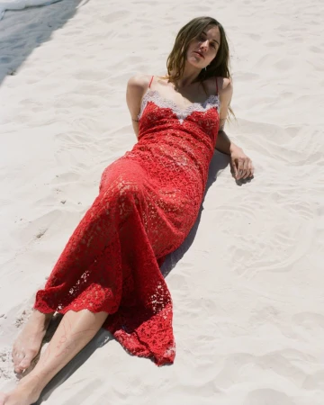 Model reclining on white sand wearing a red lace slip dress with pale lace trim for Gabriela Hearst.