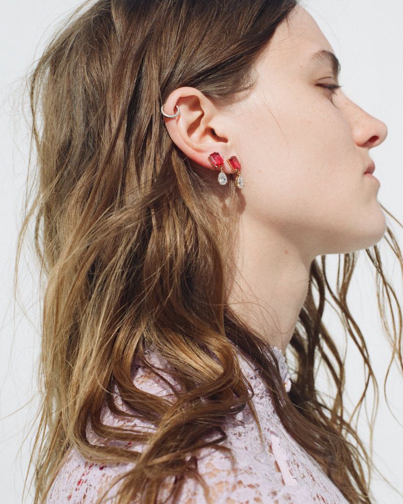 Profile view of a model with brown hair wearing red drop earrings and a high-neck pink lace top against a white backdrop.