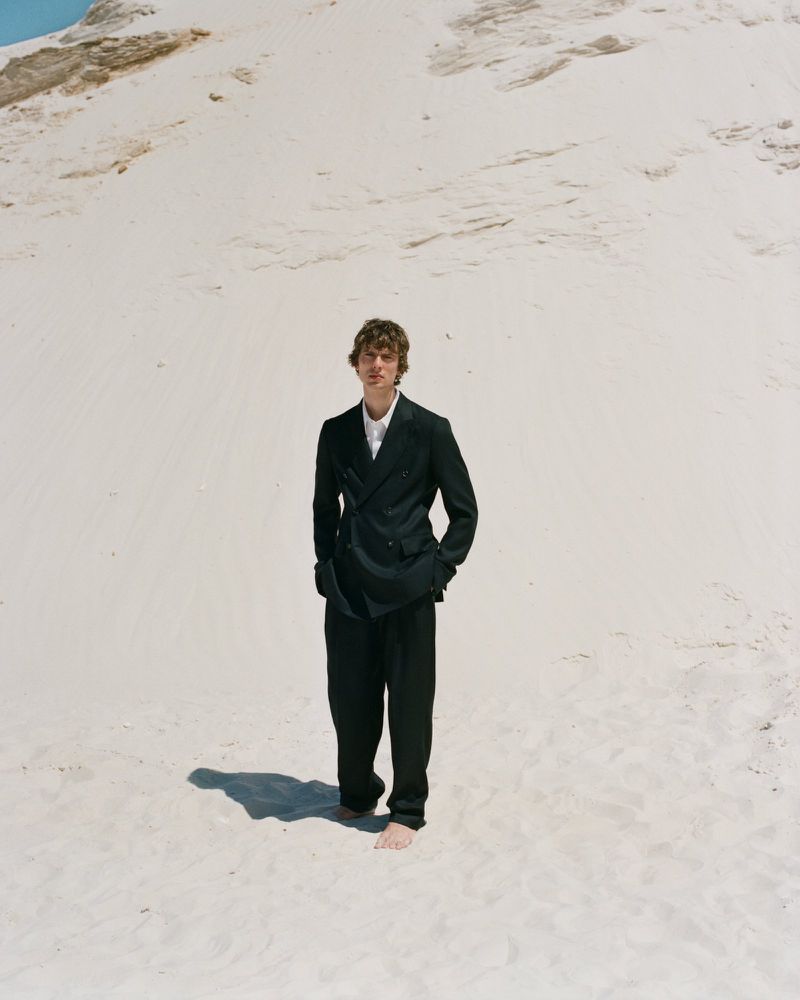 Model Leon Dame standing barefoot in a black double-breasted suit against a bright white sand dune.
