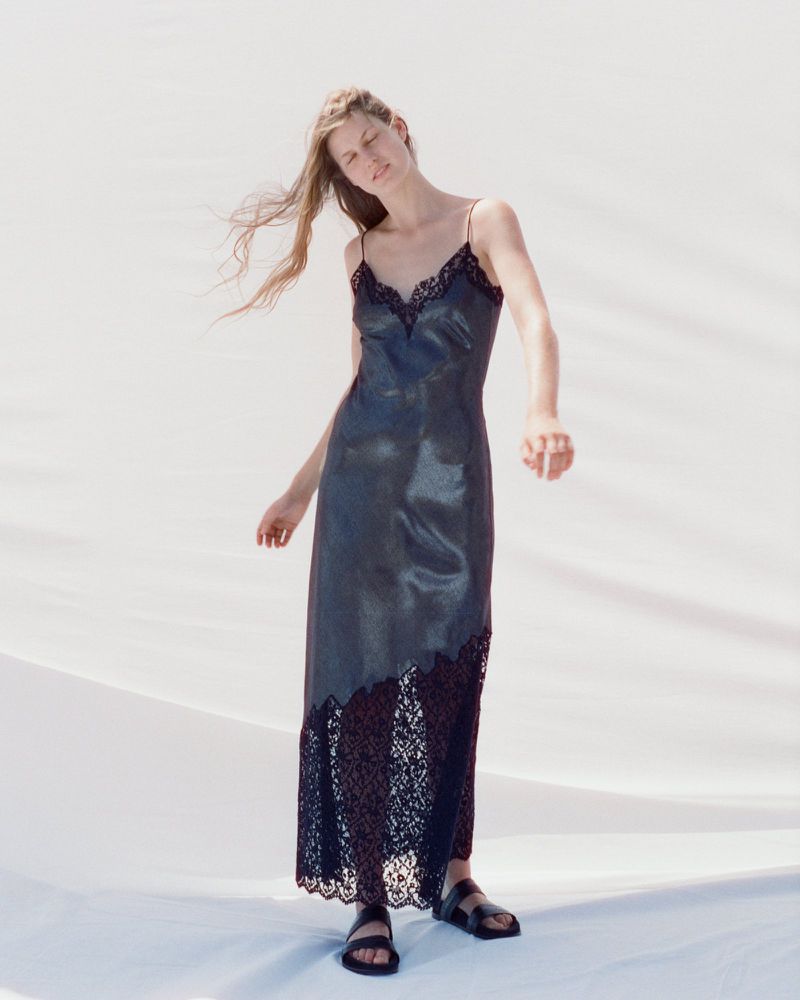 Model wearing a black lace-trimmed slip dress and black sandals standing against a bright white backdrop in natural daylight.
