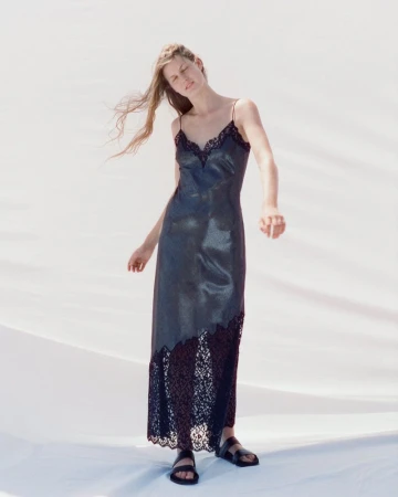 Model wearing a black lace-trimmed slip dress and black sandals standing against a bright white backdrop in natural daylight.