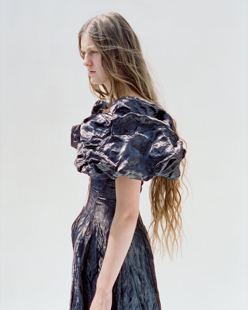 Model with long wavy hair wearing a sculptural dark textured dress against a white background