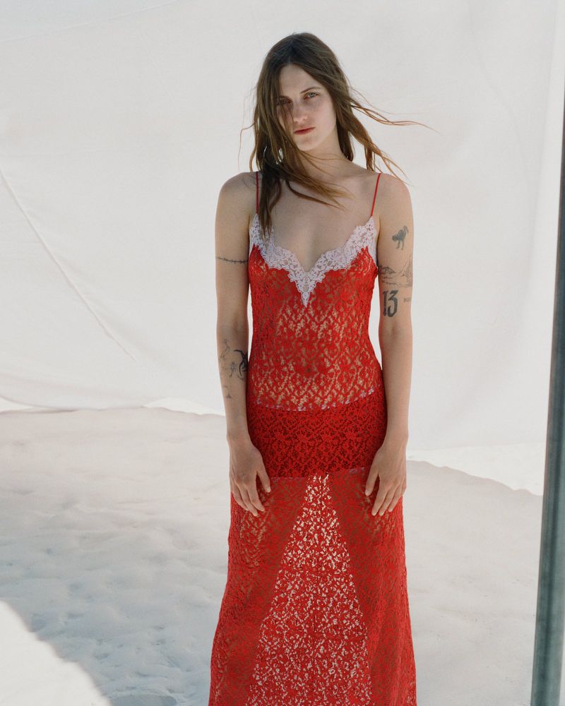 Model wearing a sheer red lace slip dress with white trim standing outdoors against a draped white fabric backdrop.