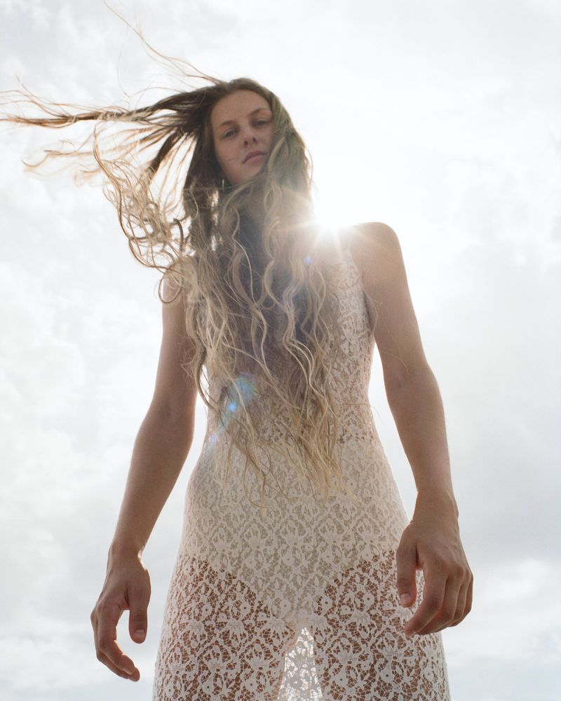Model with long windblown hair looking down into the camera wearing a sheer cream lace dress against a bright backlit sky.