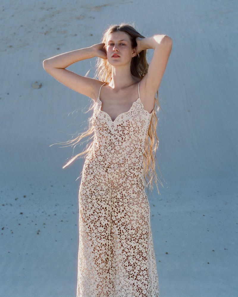 Model with long hair posing with hands behind head wearing a cream lace crochet slip dress against a pale sand dune background.