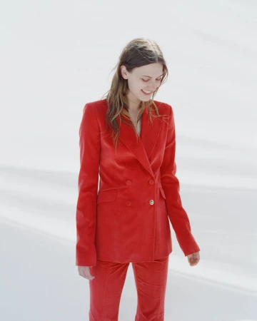 Woman smiling downward wearing a vibrant red double-breasted pantsuit against a bright white background.