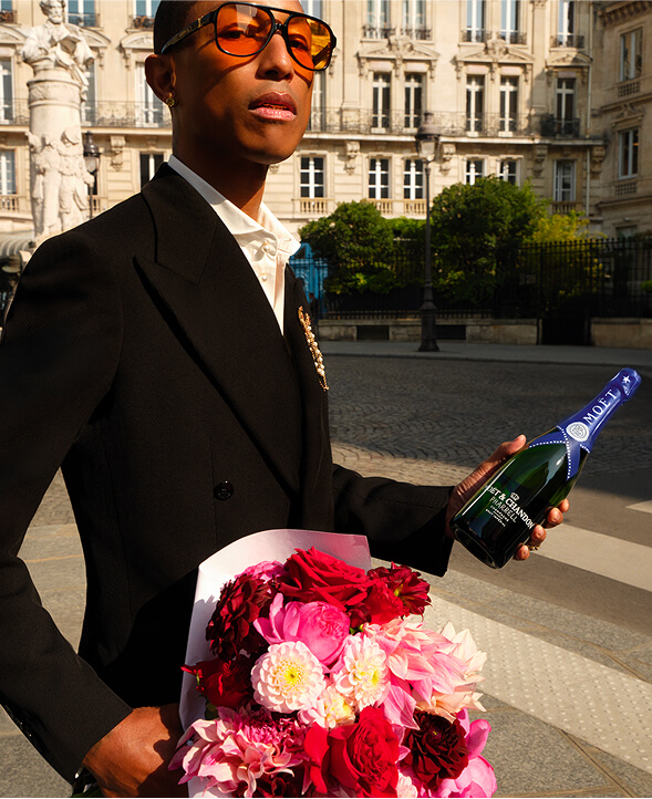 Pharrell Williams wearing amber sunglasses and a black suit jacket, holding a bouquet of pink flowers and Moët & Chandon champagne.