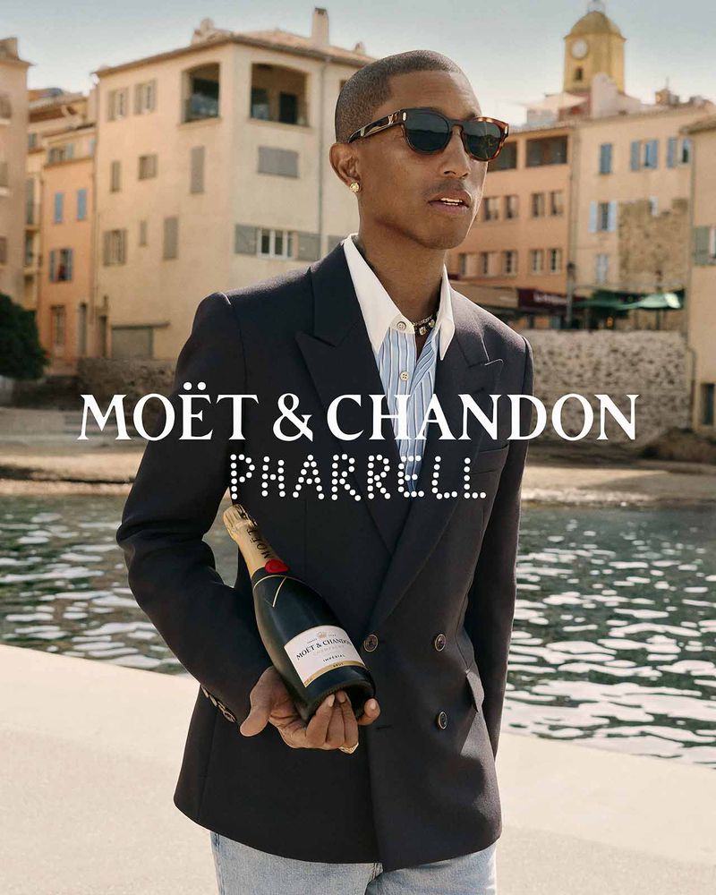 Pharrell Williams wearing a navy double-breasted blazer and sunglasses while holding a bottle of Moët & Chandon champagne.