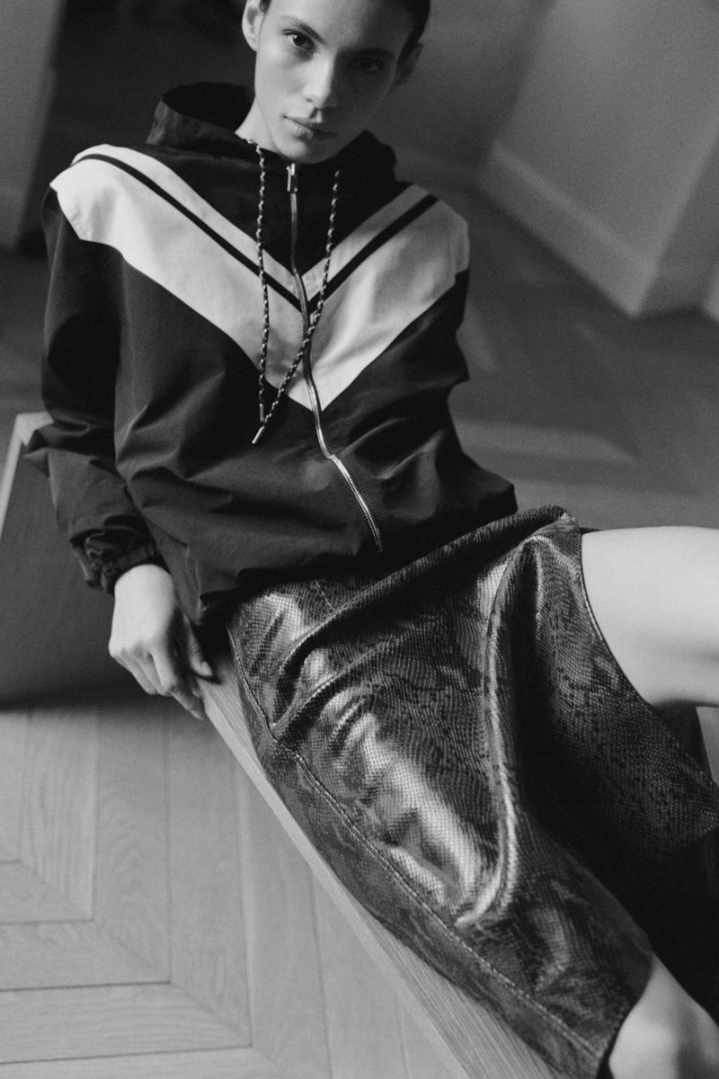 Model seated wearing a chevron zip-up windbreaker and textured snakeskin skirt on parquet flooring for Wales Bonner.