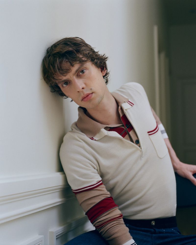Model Leon Dame leaning against a white wainscoted wall wearing a cream knit polo shirt with red stripes and navy trousers.