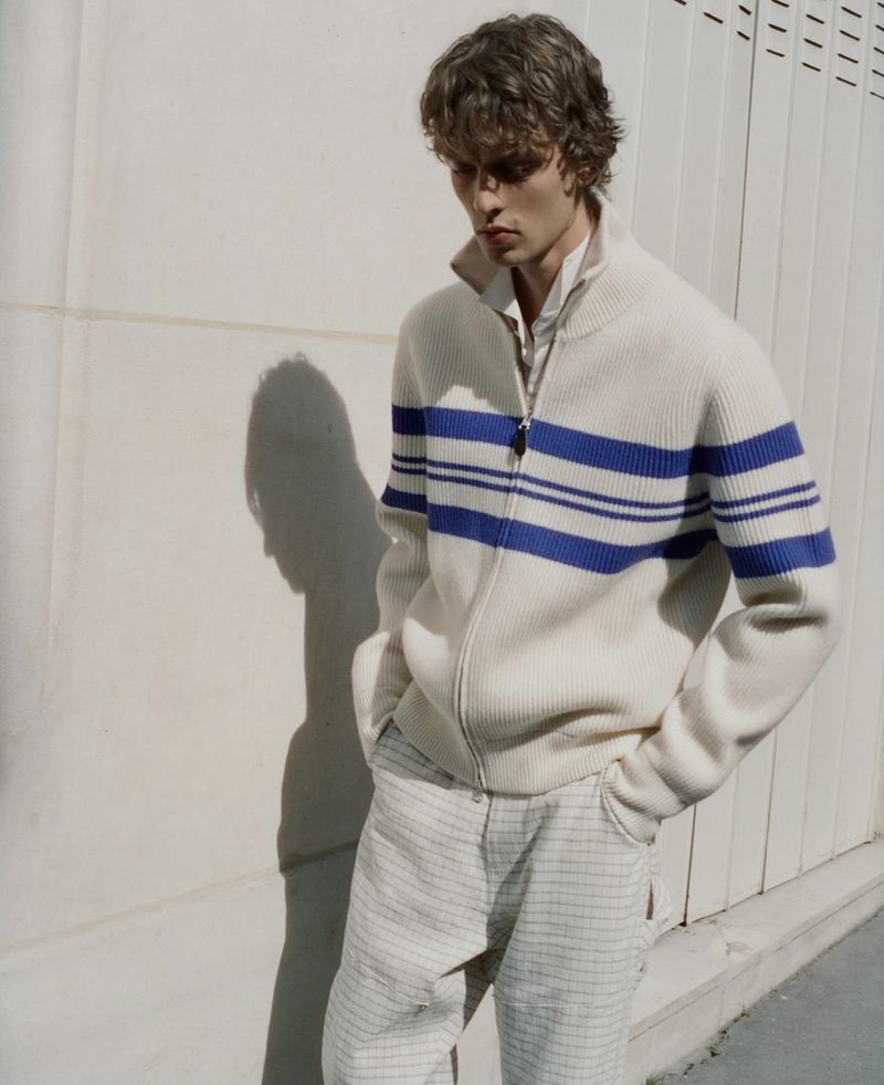 Model Leon Dame in an off-white zip-up striped knit sweater and grid pants standing against a pale sunlit wall.