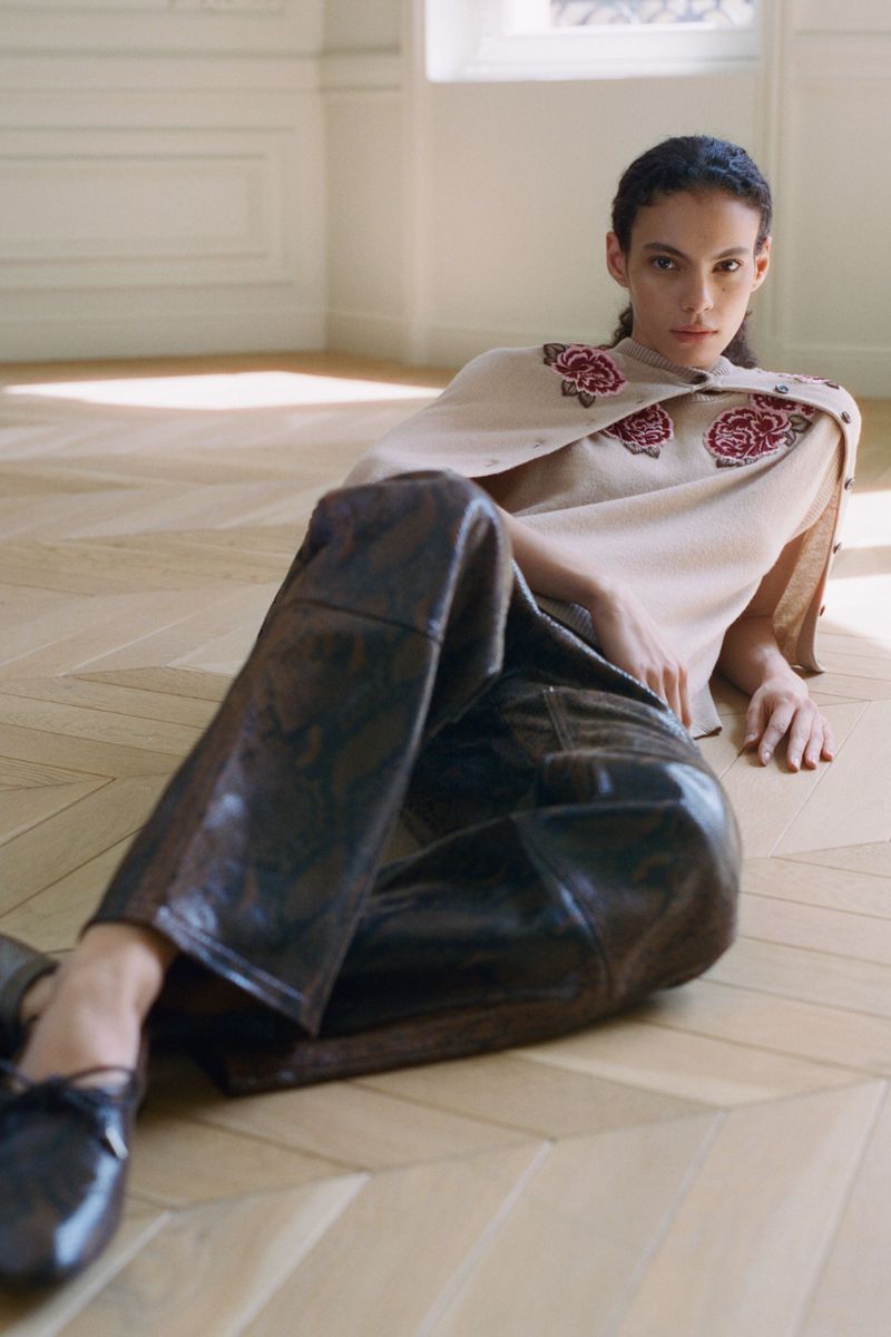 Model reclining on a chevron parquet floor wearing a light embroidered knit top and dark brown leather trousers.