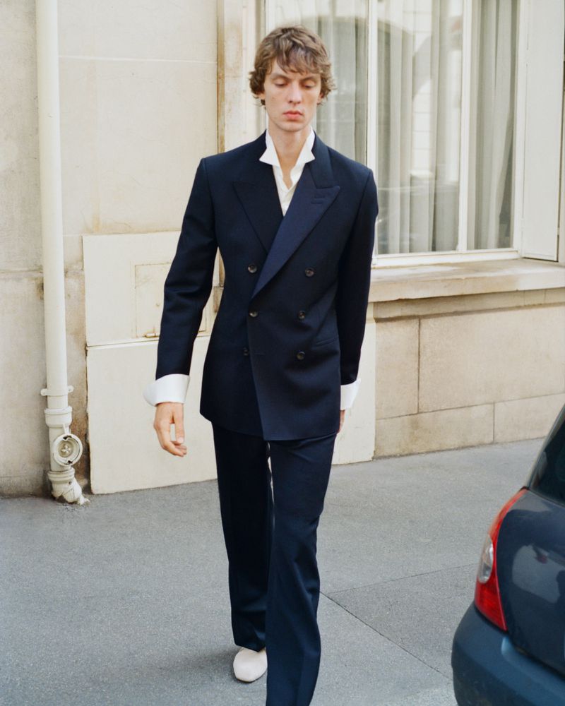 Model Leon Dame walking in a dark navy double-breasted suit and open white shirt outdoors for Wales Bonner.