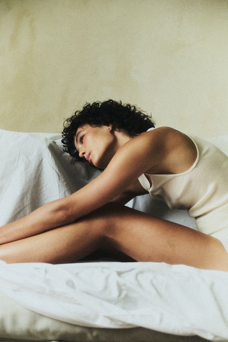 Model with short dark curly hair wearing an off-white ribbed tank top leaning forward on white bedsheets against a textured wall.