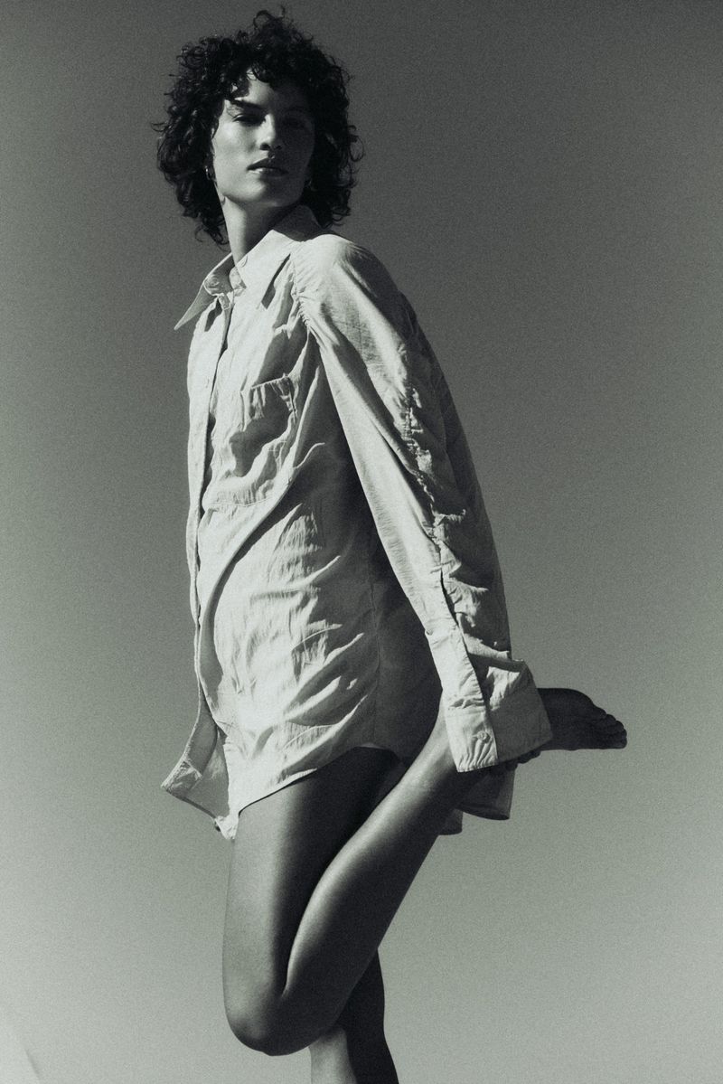 Model with dark curly hair wearing an oversized button-down shirt against a clear sky background