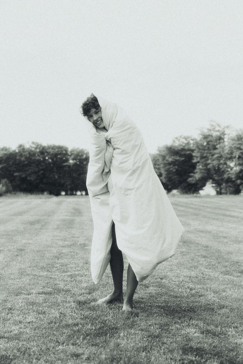 Maja Zimnoch wrapped in a large white duvet standing barefoot on a grassy field in black and white.