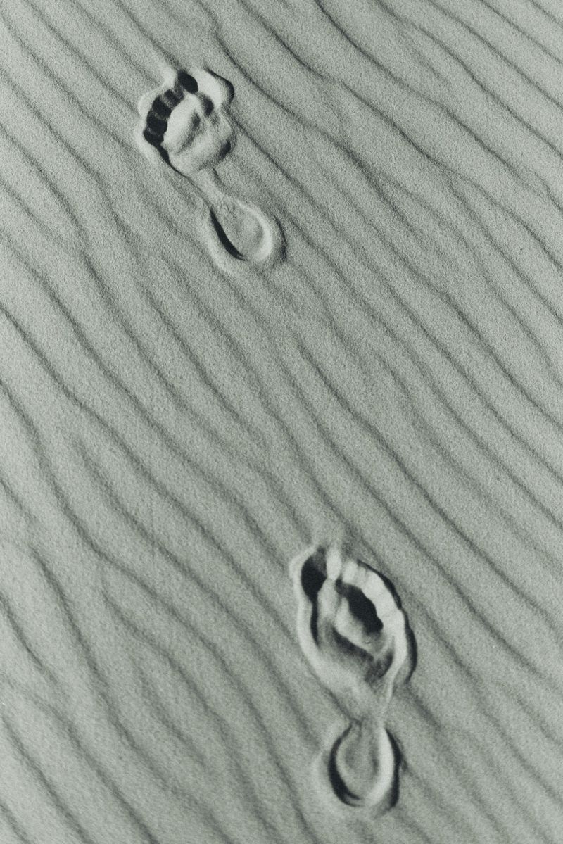 Overhead shot of two distinct barefoot footprints pressed into textured, diagonally rippled sand in black and white.
