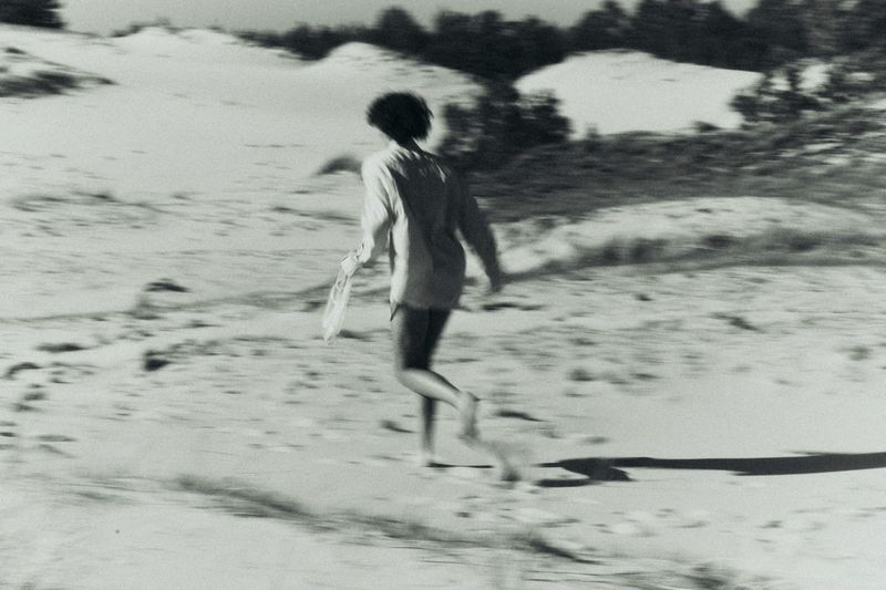 Monochrome motion shot of a woman in an oversized light shirt running across sandy dunes with dunes in the blurred background.