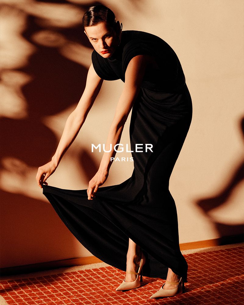 Model Saskia de Brauw in a long black dress holding the hem against a patterned wall for Mugler