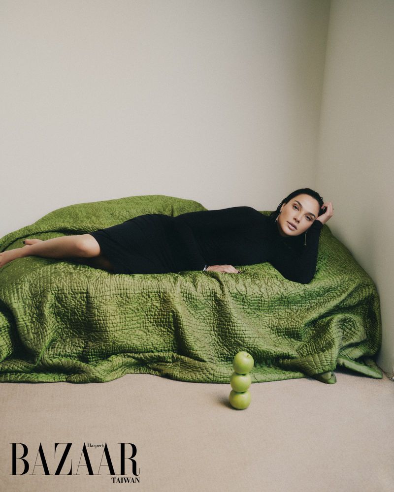 Gal Gadot lying on a textured green quilted daybed wearing a black long-sleeve dress with three stacked green apples on the floor.